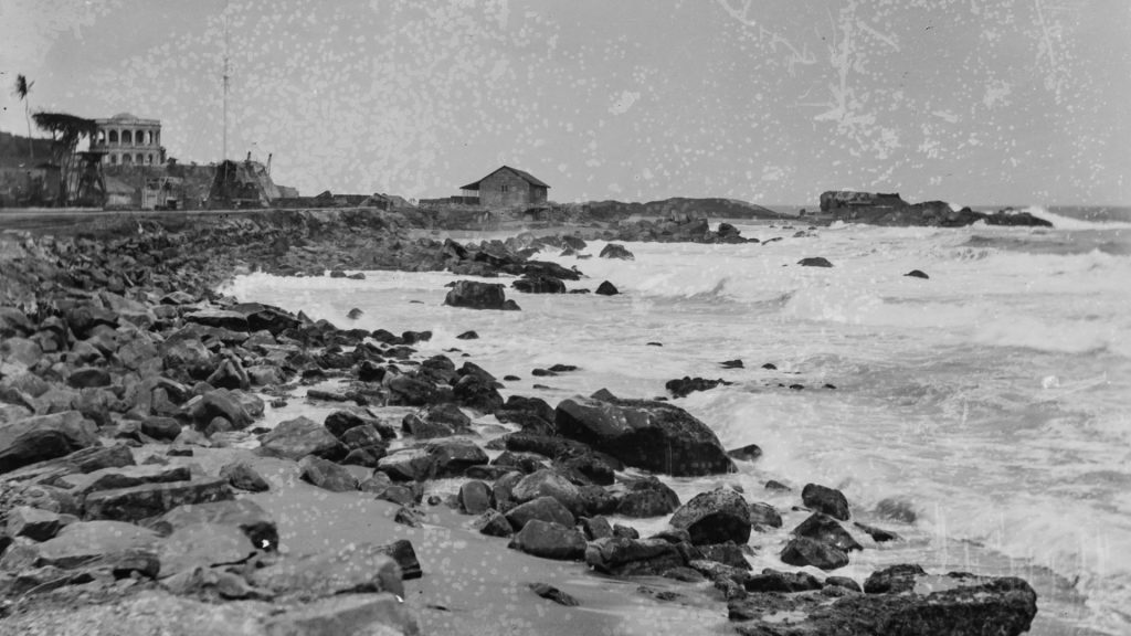 Black and white coastal scene rocky shoreline with waves a small house on a rock outcrop and ruins on the left Image