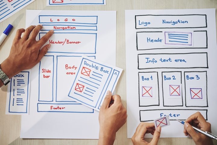 Image | how to select website layouts for different business categories | entheosweb Two people are sketching website wireframes on white paper using blue and red markers The left sheet shows blocks labeled logo navigation headerbanner body area slide bar and footer The right sheet shows logo navigation header info text area three content boxes box 1box 3 and a footer A hand is placing a printed card with a layout sketch on the table Image